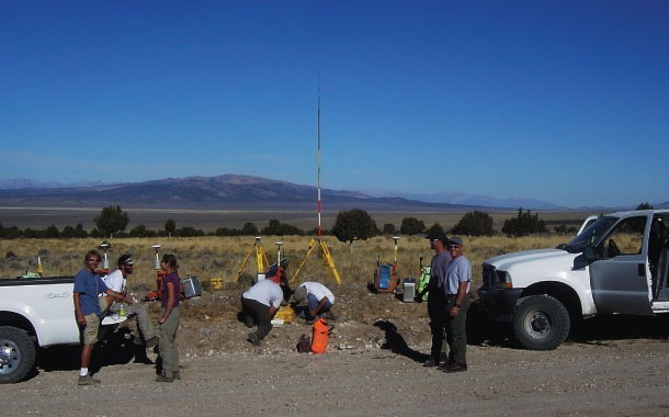 Field researchers conduct a geological survey in a remote area, with equipment set up nearby and mountainous terrain in the background.